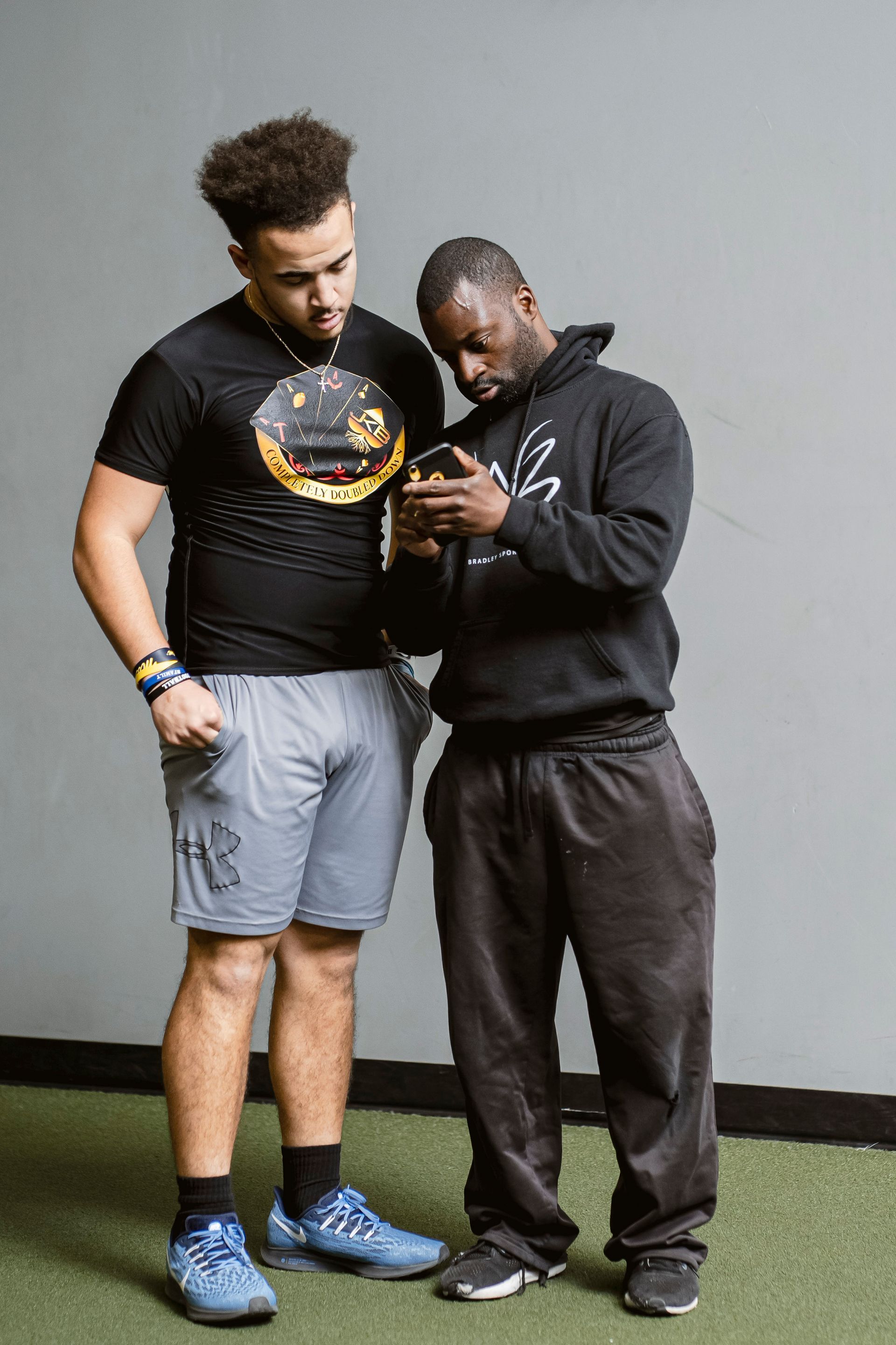 Two men standing indoors, looking down at a phone, one pointing at the screen.