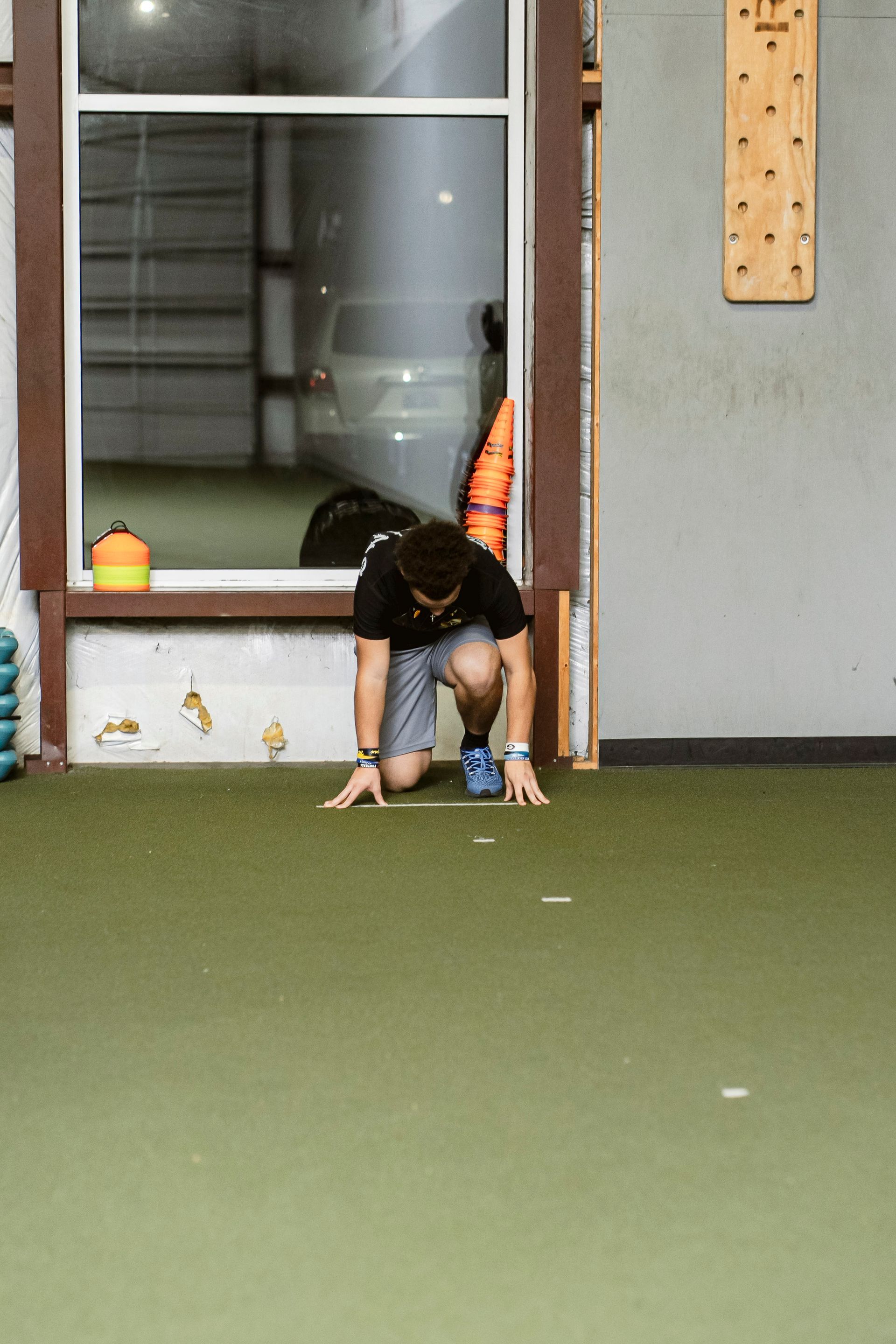 Person crouching on indoor turf near a window, reaching toward a small blue ball with a toy nearby.