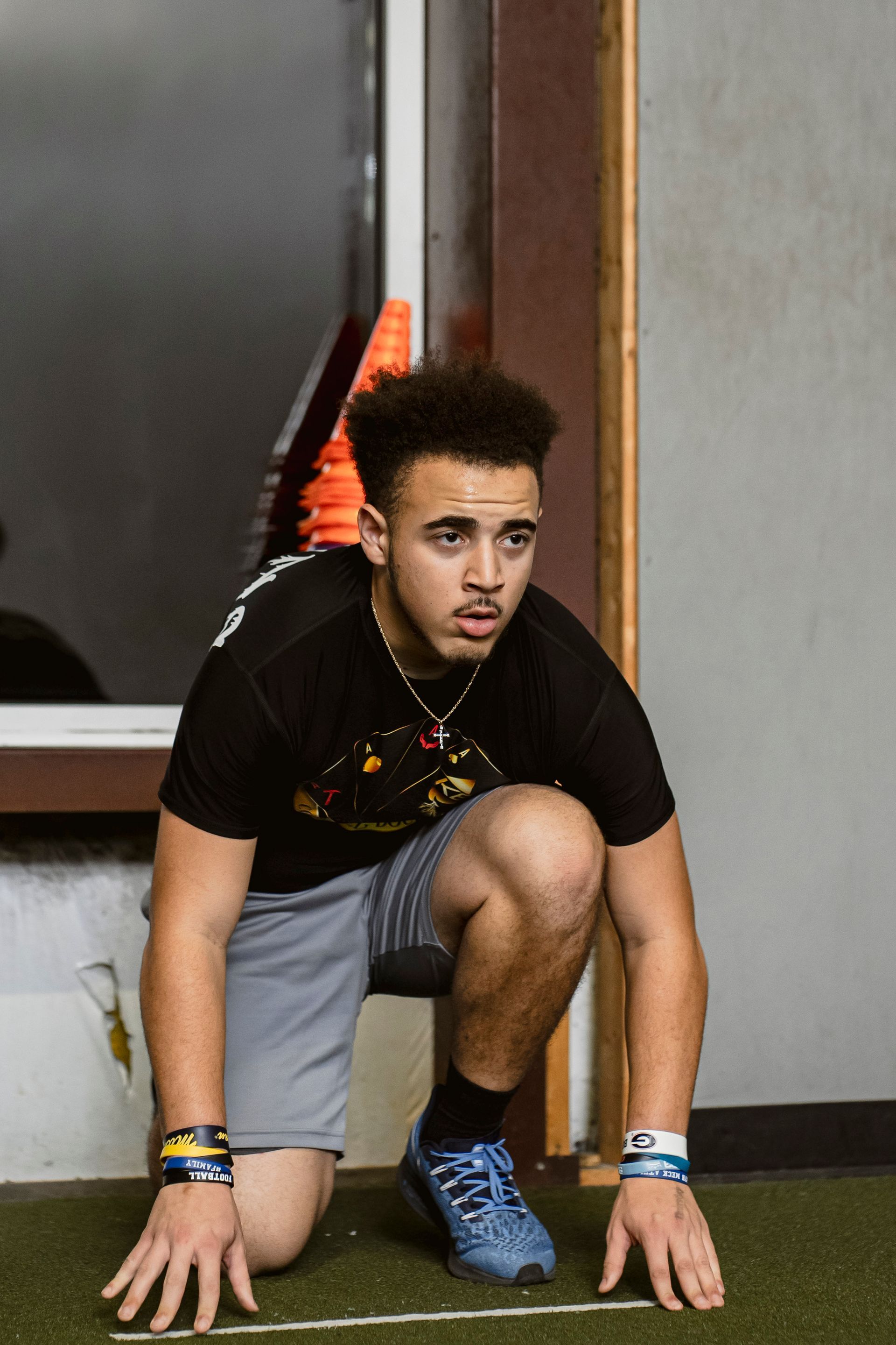 Athlete crouching at the starting line indoors, hands on the floor, wearing a black shirt and gray shorts.
