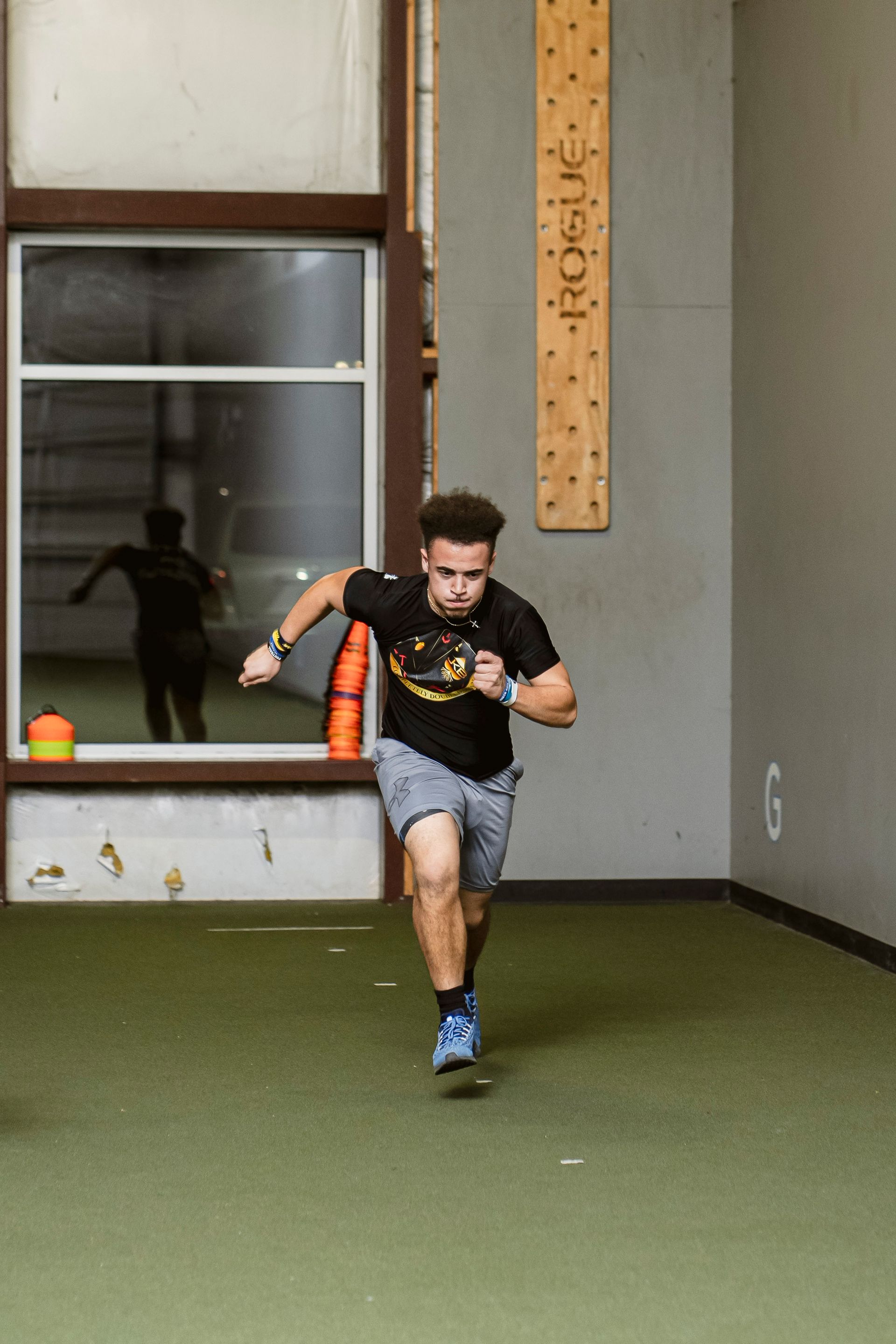 Athlete sprinting indoors on turf, holding a baton, with orange cones and a windowed wall behind him.