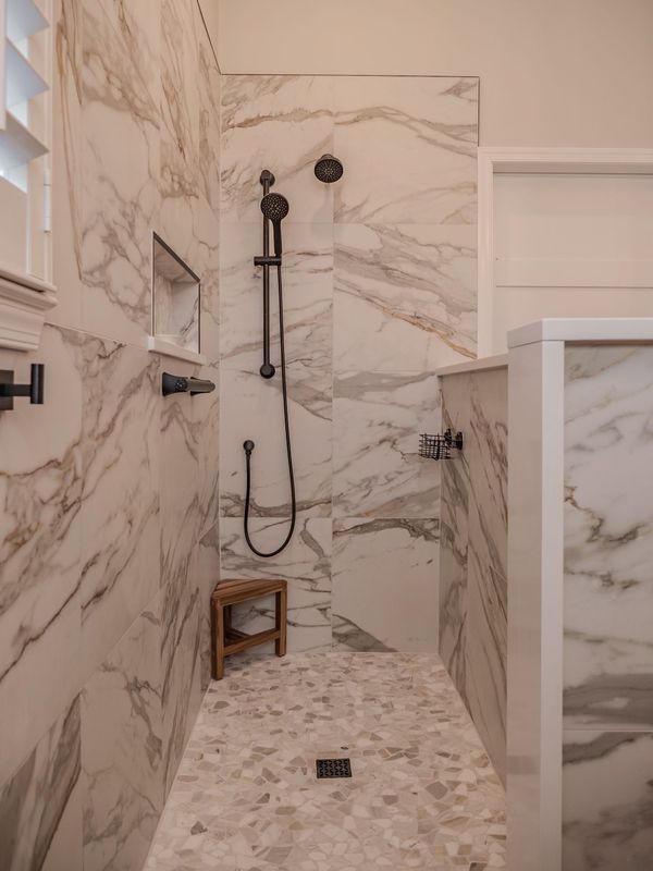 Marble-tiled shower with a pebble floor, black fixtures, and a wooden stool.