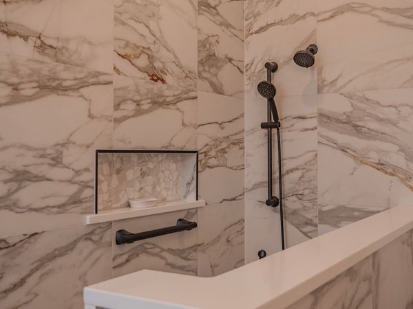 Bathroom shower with marble-like tile, dark shower fixtures, and a built-in shelf with a grab bar.