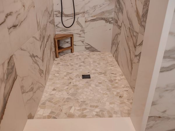 Shower with marble-patterned walls and a mosaic floor, featuring a small wooden stool and a black showerhead.