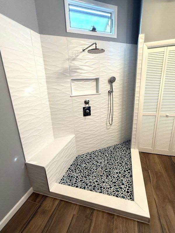 Bathroom with white wavy tile shower, bench, and pebble floor; small window, silver fixtures, and wood-look floor.