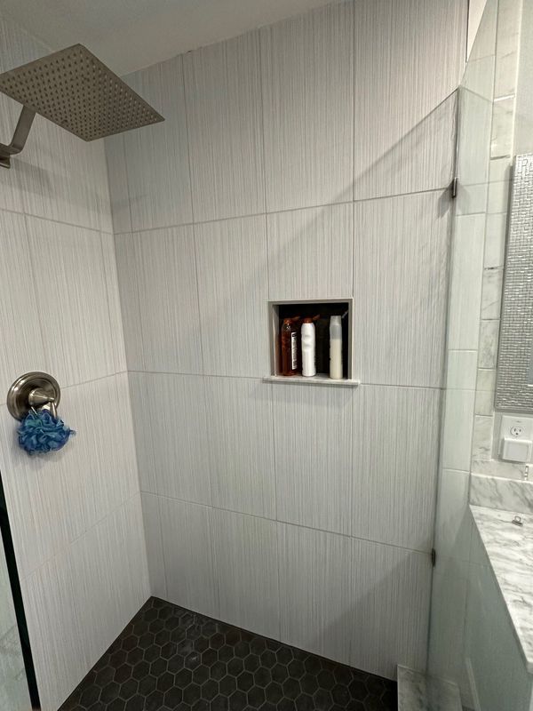 Shower with white vertical-striped tiles, a recessed shelf with bottles, a dark hexagonal floor, and a silver shower head.