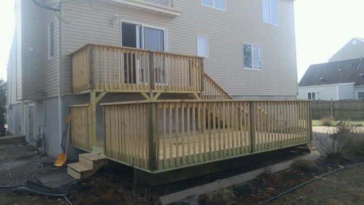 Two-tiered wooden deck attached to a light-colored house. Stairs lead up to the top level. The yard is bare.