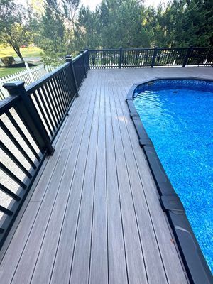 Gray composite deck with black railing surrounds a blue swimming pool. Green trees in background.
