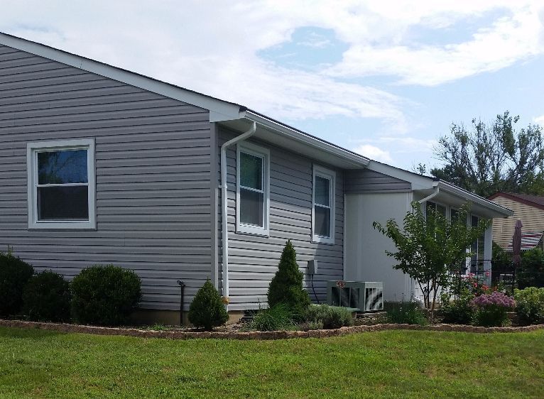 Gray vinyl-sided house with white window frames and a landscaped front yard on a sunny day.