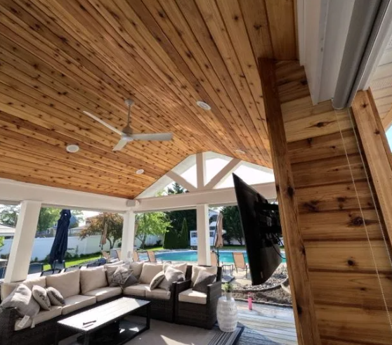 Outdoor covered patio with wood ceiling, fan, and mounted TV. Features seating, a pool, and white support beams.