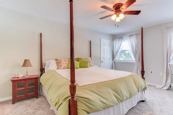 Bedroom with four-poster bed, green bedding, nightstand with lamp, window with curtains, and ceiling fan.