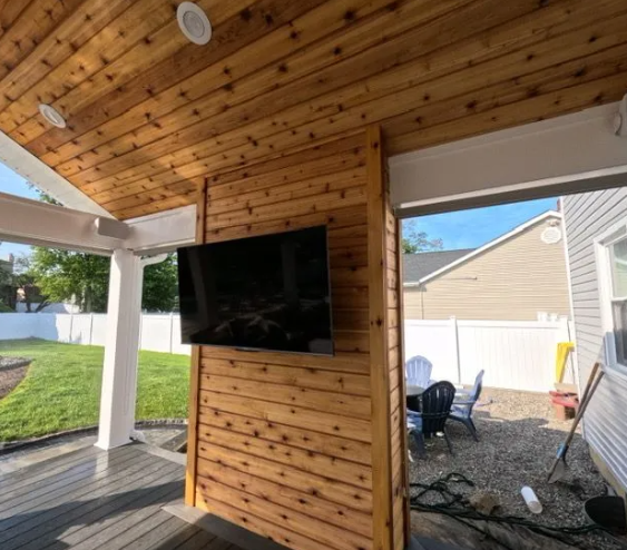 Outdoor patio with wooden ceiling and wall-mounted TV. Lawn, fence, and patio furniture visible.