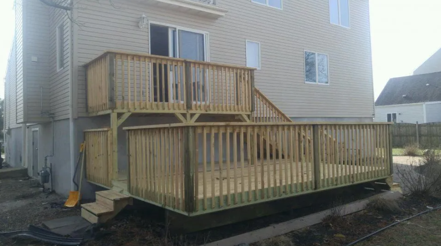 Two-level wooden deck attached to a light beige house. Sunlight.