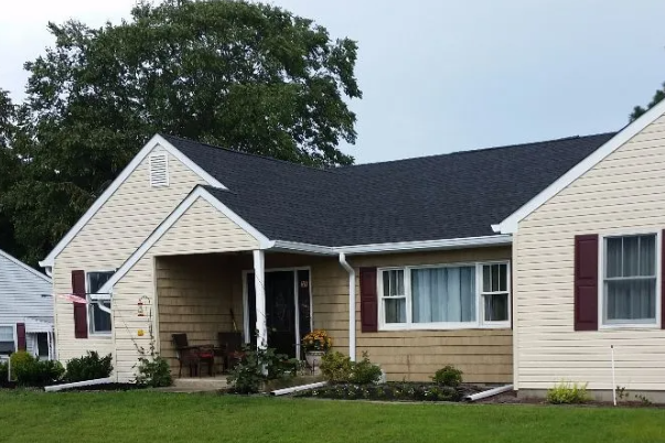 Beige ranch house with dark roof and red shutters, green lawn under overcast sky.