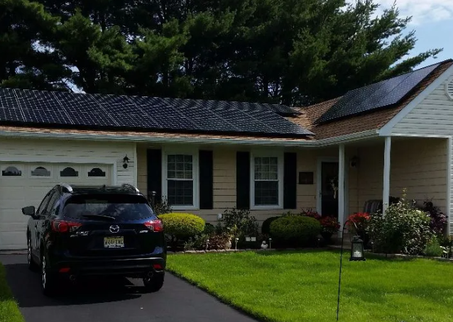 Solar panels on a tan roof of a one-story home with a black car in driveway. Green lawn and bushes in front.