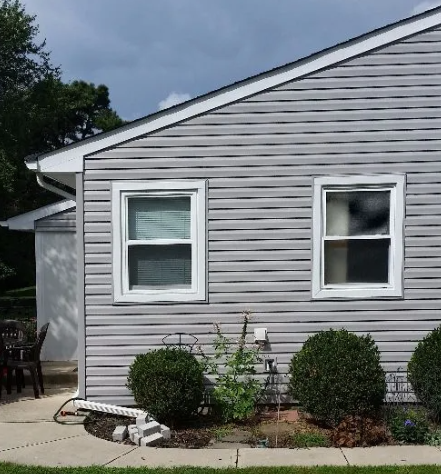 Gray house with white-framed windows, surrounded by bushes, and a small yard on a cloudy day.
