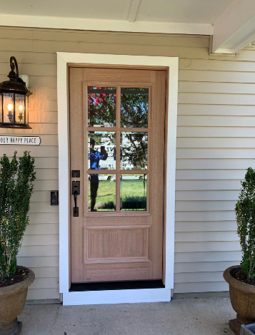 Wooden front door with glass panels, flanked by potted plants, beneath a porch light on a tan house.
