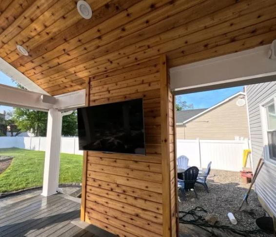 Covered outdoor patio with a TV mounted on a wooden wall. Cedar ceiling, white columns, and backyard view.