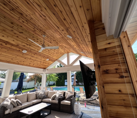 Wooden outdoor covered patio with seating, ceiling fan, and television, overlooking a pool.
