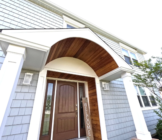Front entrance of a house with a brown door and arched wood paneling above. Shingle siding.