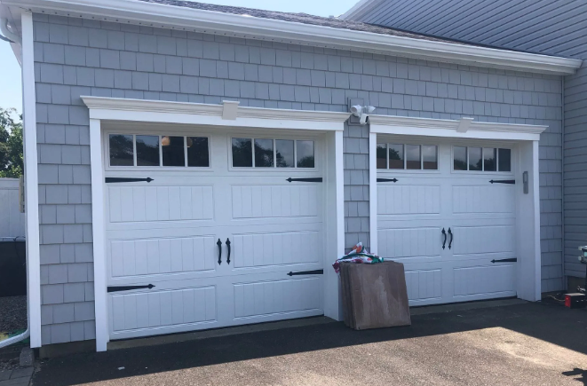 Two white garage doors with black hardware, set in a blue-gray shingle siding wall.