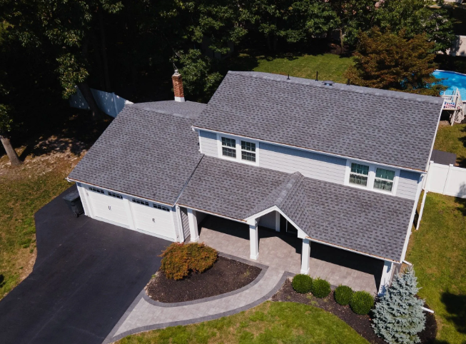 Aerial view of a gray-roofed house with white garage doors and a paved driveway, surrounded by greenery.