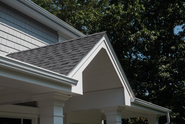 Gray-shingled roof and white trim of a house with a porch and trees in the background.