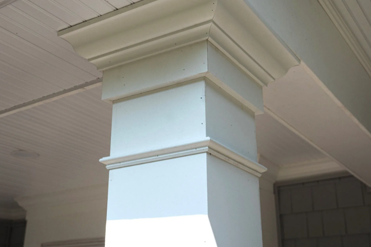 Box-shaped, white painted porch column with decorative molding at the top and bottom, next to a white ceiling.