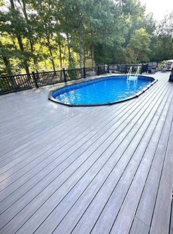 Gray composite deck surrounds a blue oval pool.  Black fence and trees are in the background.