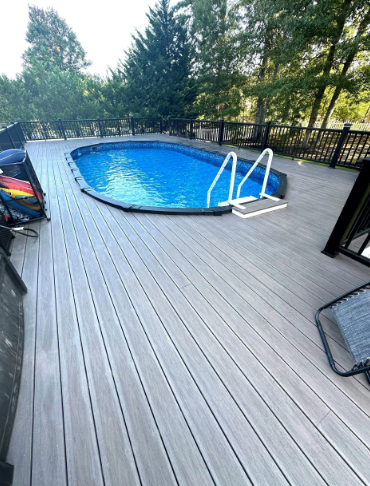 Grey composite deck surrounds an oval above-ground pool with ladder. Black railing and trees in the background.