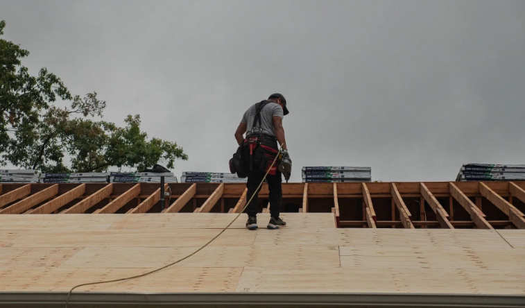 Roofer on partially constructed roof under a cloudy sky, wearing safety harness and tool belt.