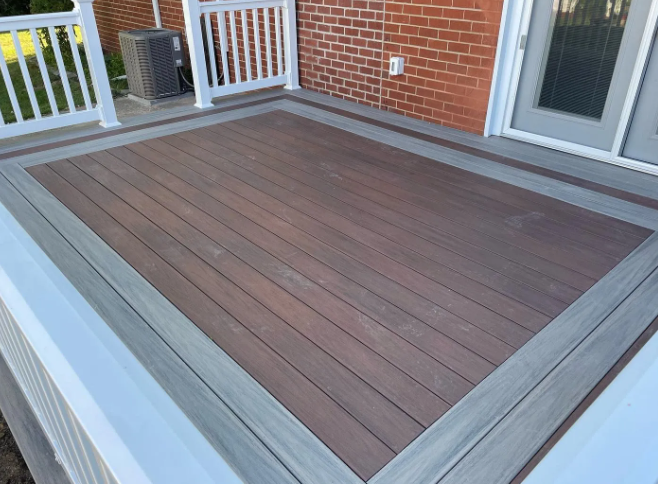 Composite deck with a brown interior and gray border, enclosed by white railings, against a brick wall.