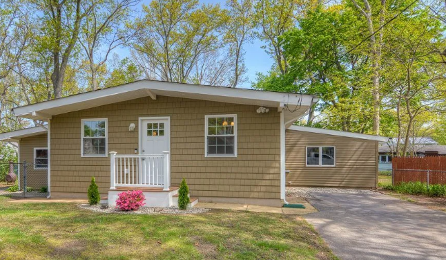 Tan bungalow with white trim, small porch, and driveway, surrounded by trees.