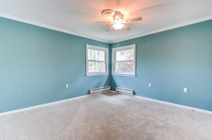 Empty room with light blue walls, two windows in the corner, and beige carpet.