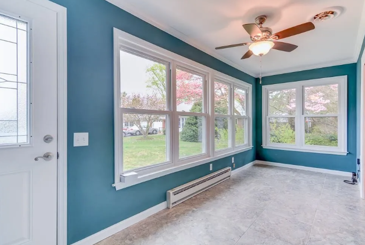Sunroom with blue walls, large windows, ceiling fan, and tiled floor, overlooking a yard.