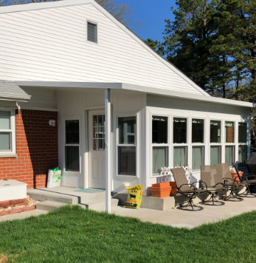 White house with a brick side and a porch with chairs on concrete next to grass under blue sky.
