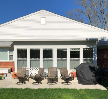 Patio with white siding, windows, and chairs on a concrete slab, with a covered grill and green grass.