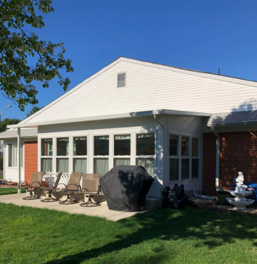 Exterior view of a white-sided house with a patio, lawn, and sunroom with multiple windows under a blue sky.