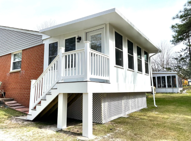 White sunroom with stairs attached to a red brick house; latticework base, door, and windows.