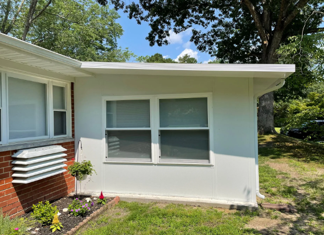 White-walled building with two windows and a brick section, surrounded by grass and trees under a blue sky.