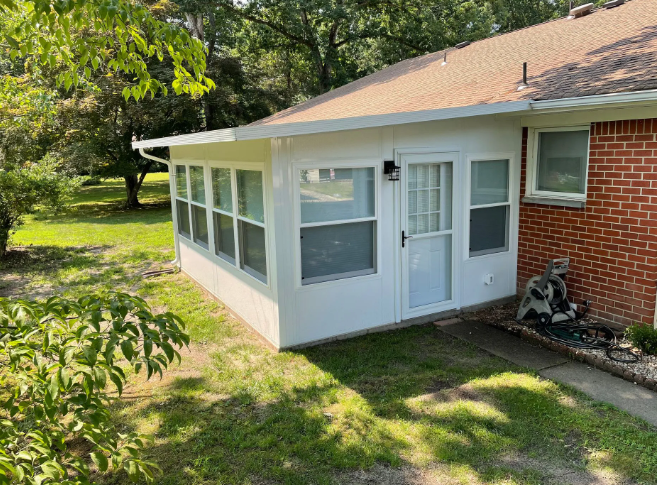 White sunroom attached to a brick building, featuring windows, a door, and a brown roof, surrounded by grass and trees.