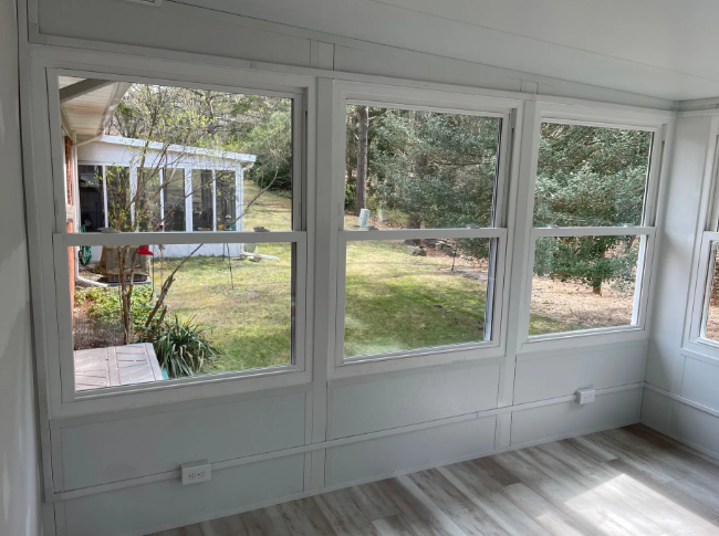 Interior view of a sunroom with three large windows overlooking a backyard and a small white structure.