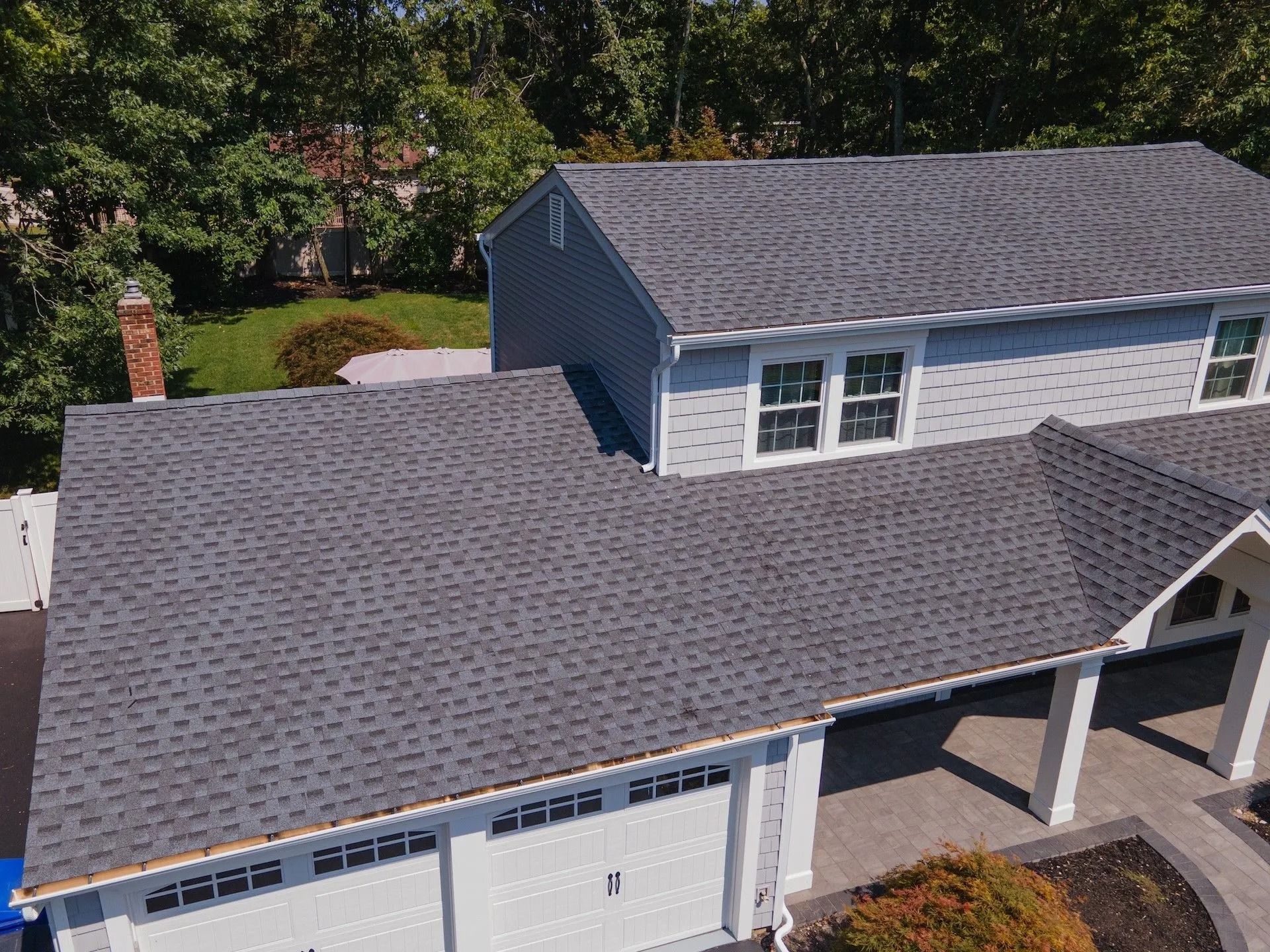 Gray-shingled roof on a two-story house with a white garage. Trees in the background, blue sky.