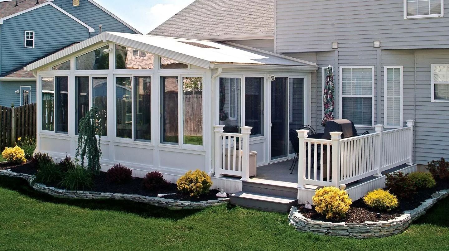 White sunroom with windows attached to a house with a deck; green lawn and flowerbeds surround it.