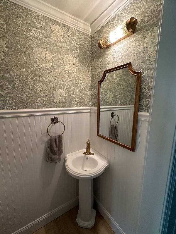 Small bathroom with floral wallpaper, pedestal sink, gold mirror, and wall sconce.