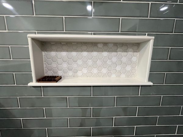 Built-in shower niche with white frame, inset with white hexagon tile, set in a tiled wall with green subway tiles.