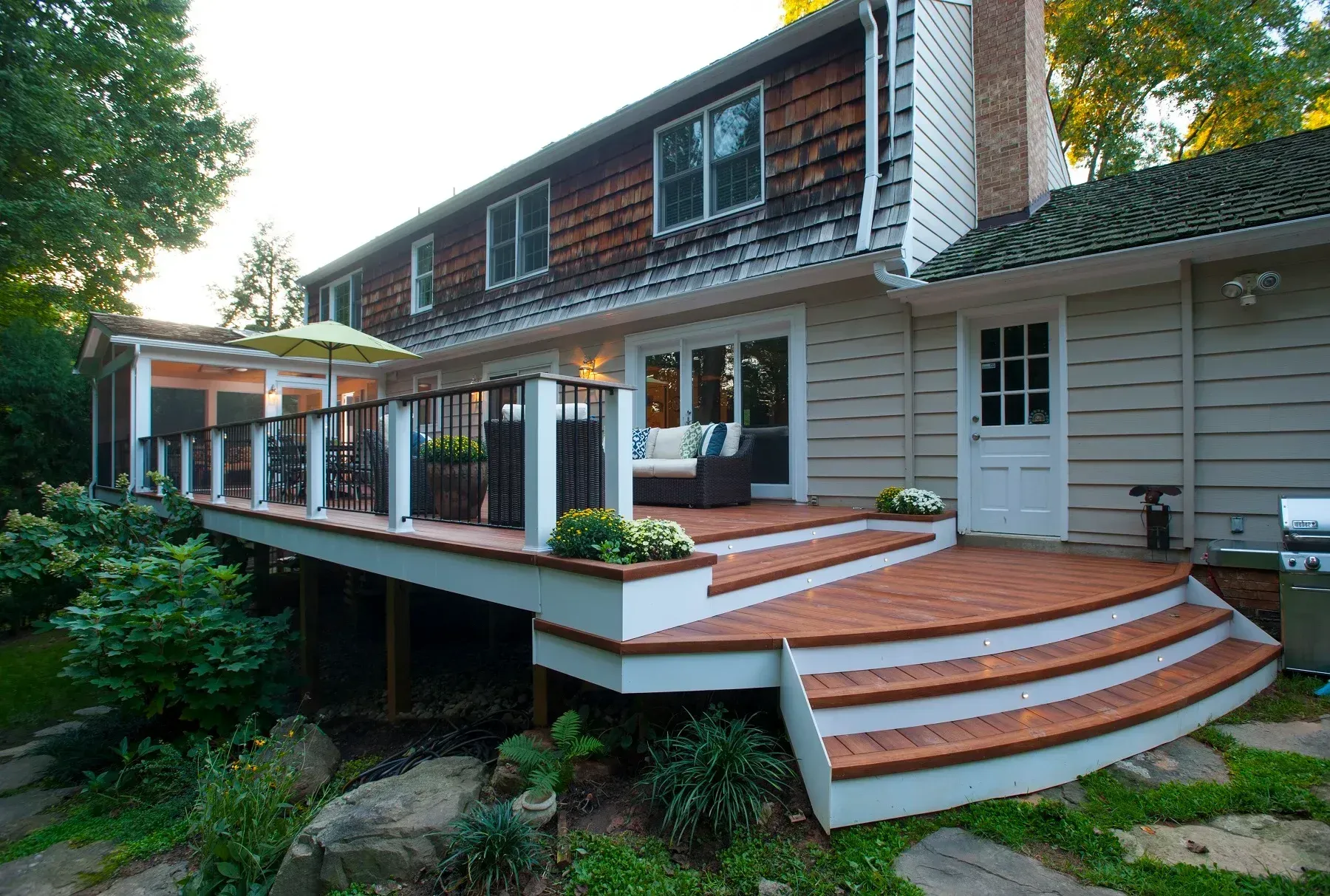 Wooden deck with steps leading to a house. Beige siding, brown roof, and greenery surround.