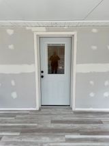 White door with glass pane, framed by white trim, set in a gray wall. Gray and white flooring.