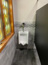 Urinal in a bathroom with gray tile walls, stained glass window, and dark stall divider.