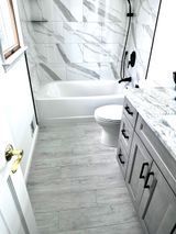 Bathroom with gray wood-look floor, white tub, marble tile, gray vanity, and black fixtures.