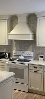 Kitchen with white cabinets, stove, and range hood. Gray backsplash. Wooden floor.